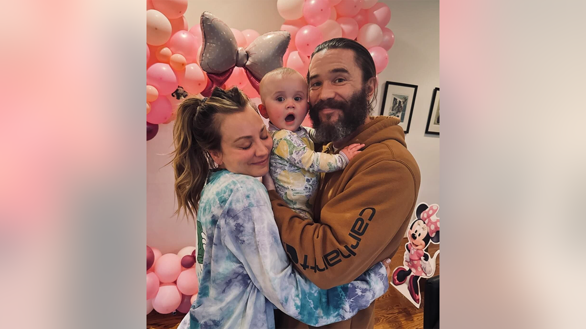 Kaley Cuoco in a tie-dye shirt hugs husband Tom Pelphrey in a burnt orange sweatshirt holding their daughter Matilda