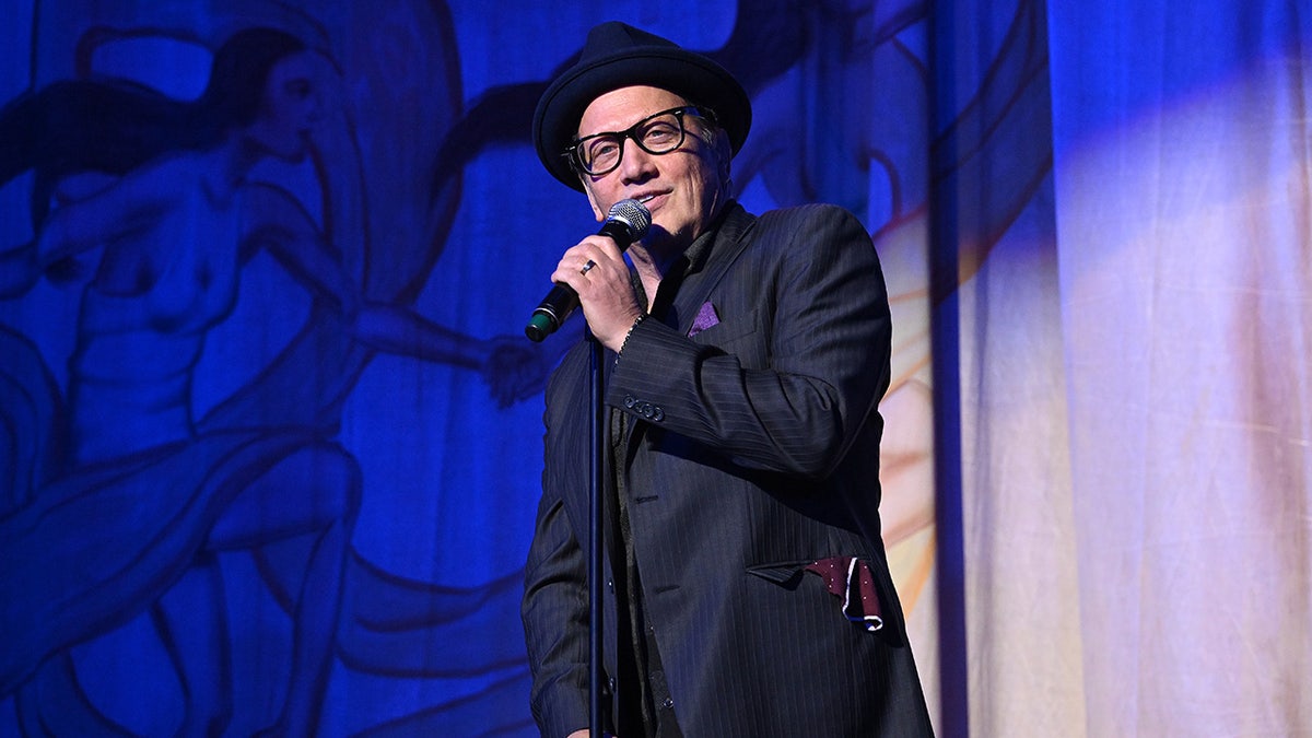 Rob Schneider in a top hat and navy suit performs on stage