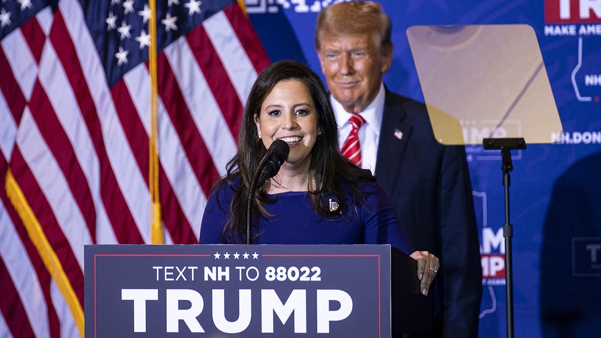 Elise Stefanik speaking at lectern with Trump behind her