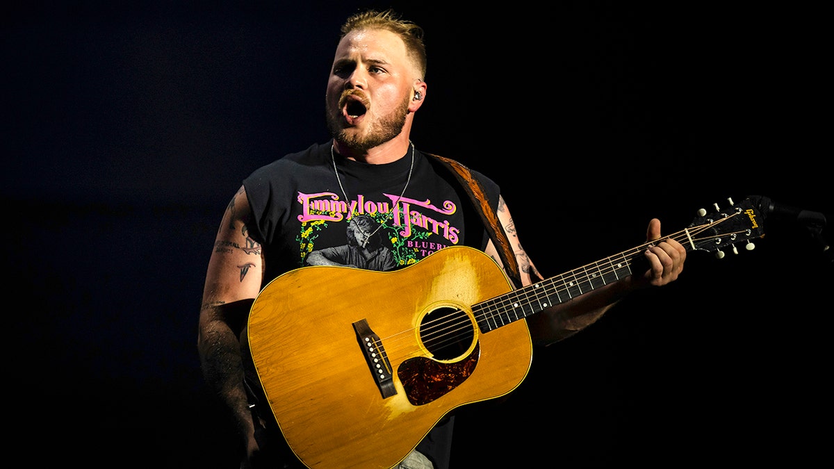 Zach Bryan in a cut off black shirt looks animated/slightly angry on stage with his mouth agape with a guitar