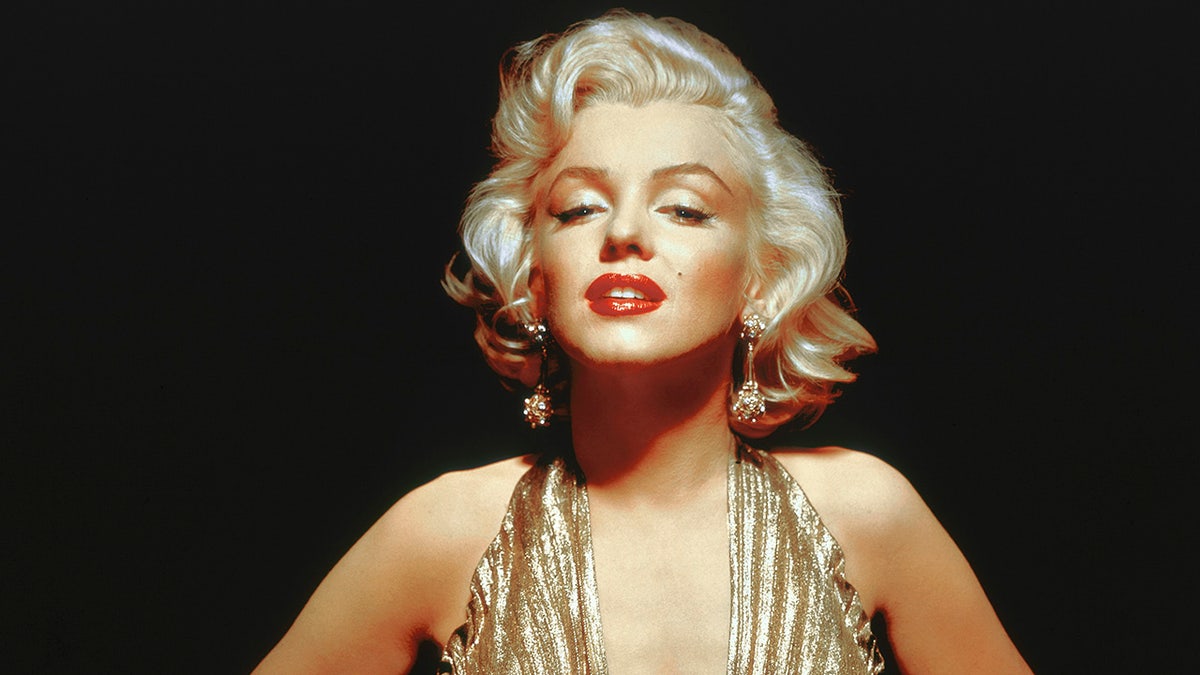 Marilyn Monroe poses seductively, leaning towards the camera wearing a gold dress