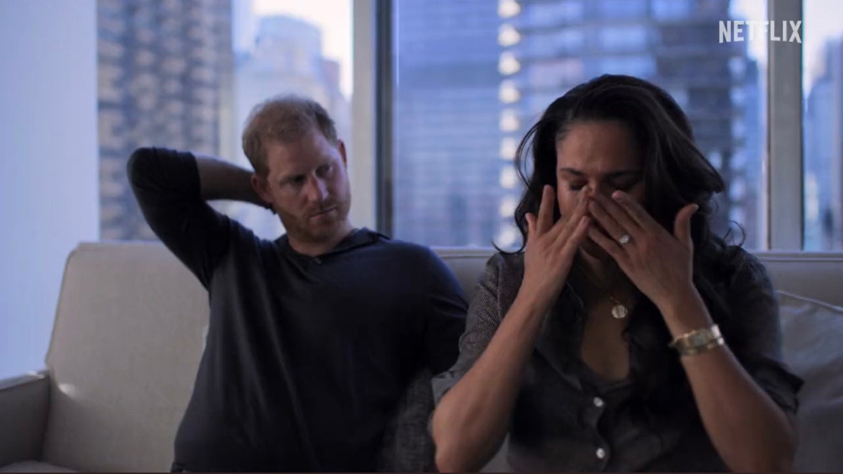 Meghan Markle cries on a couch and puts her hands to her face as Prince Harry with his arm behind his neck looks at her