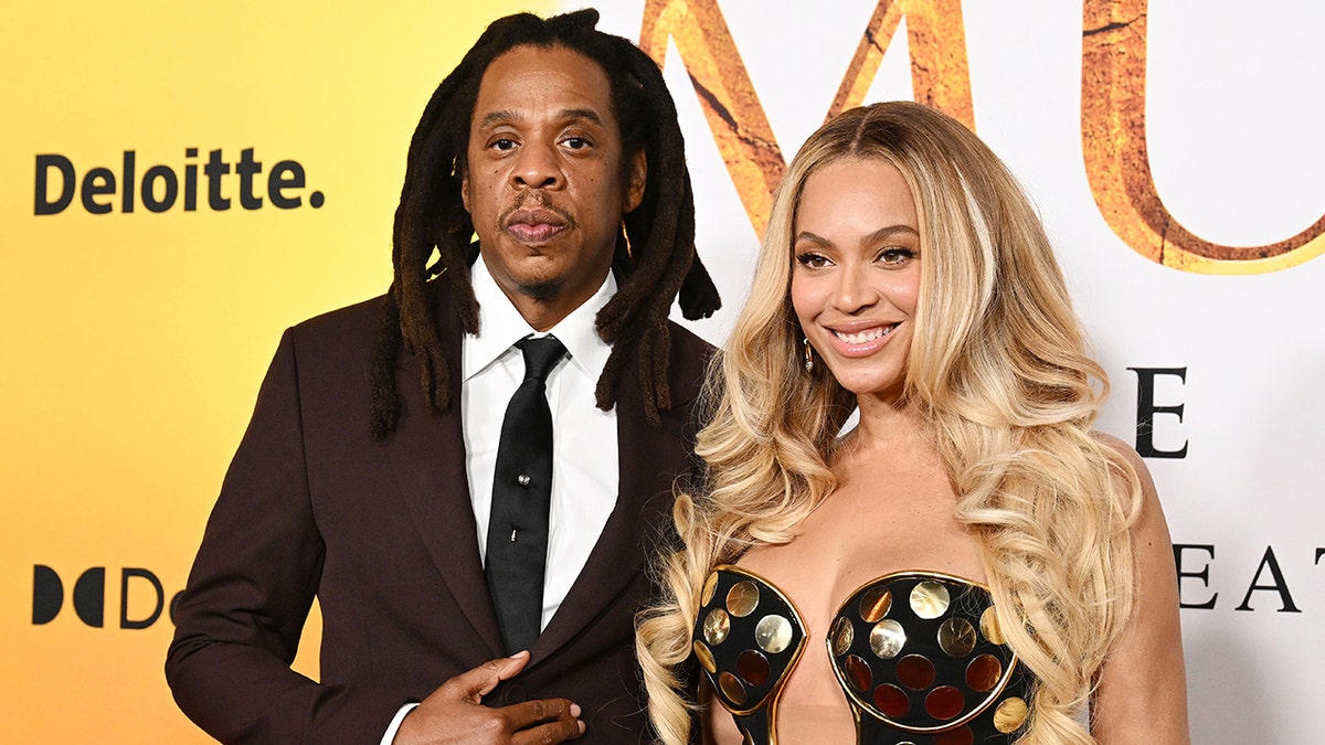 Jay-Z in a maroon jacket looks directly at the camera while wife Beyoncé in a gold circle dress smiles on the 'Mufasa: The Lion King' carpet