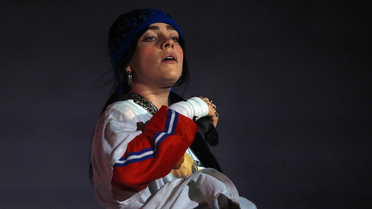 Billie Eilish in a blue beanie and multicolored long sleeve shirt performs on stage, with her head slightly tilted back