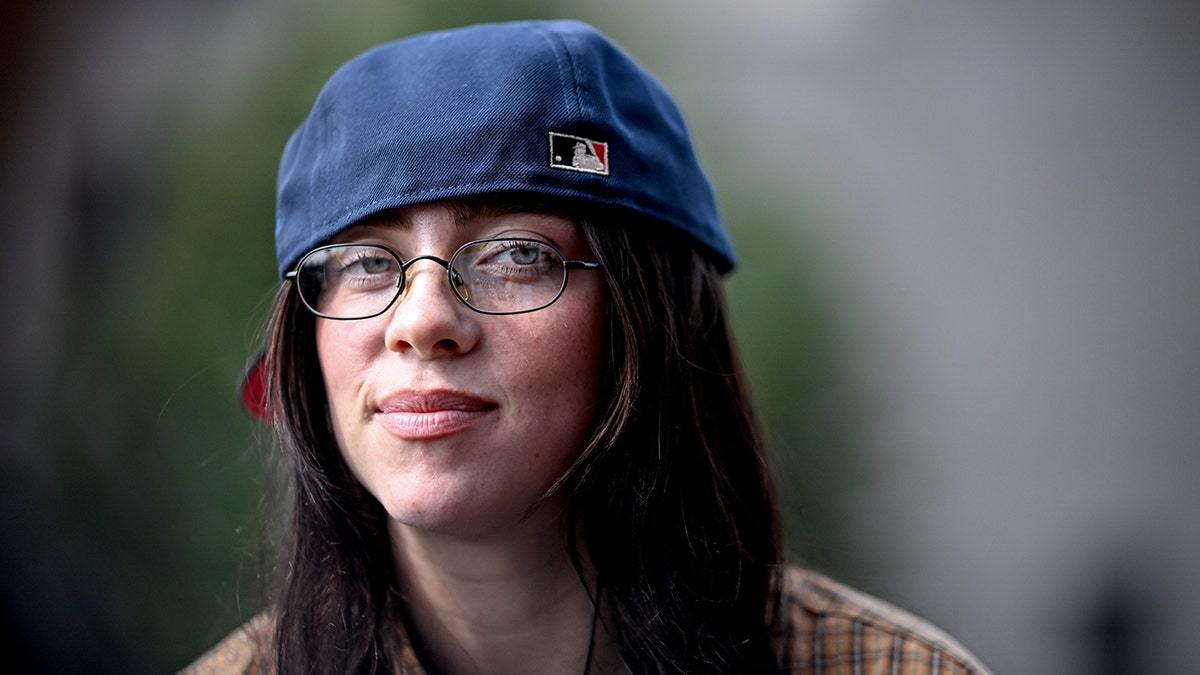 Billie Eilish in a backwards baseball hat soft smiles wearing glasses