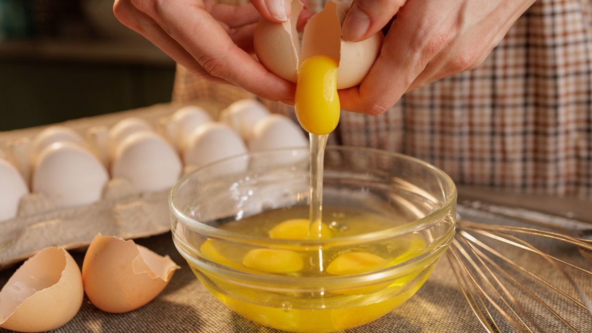 A person cracks an egg into a glass bowl, with several eggs in an egg carton in the background. The scene is set in a kitchen, showcasing the process of preparing food.