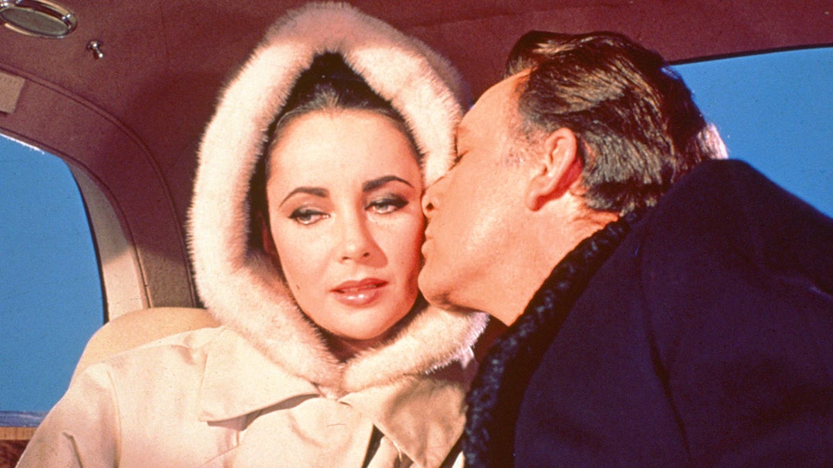 Richard Burton kissing Elizabeth Taylor on the cheek as she wears a hooded white fur coat.