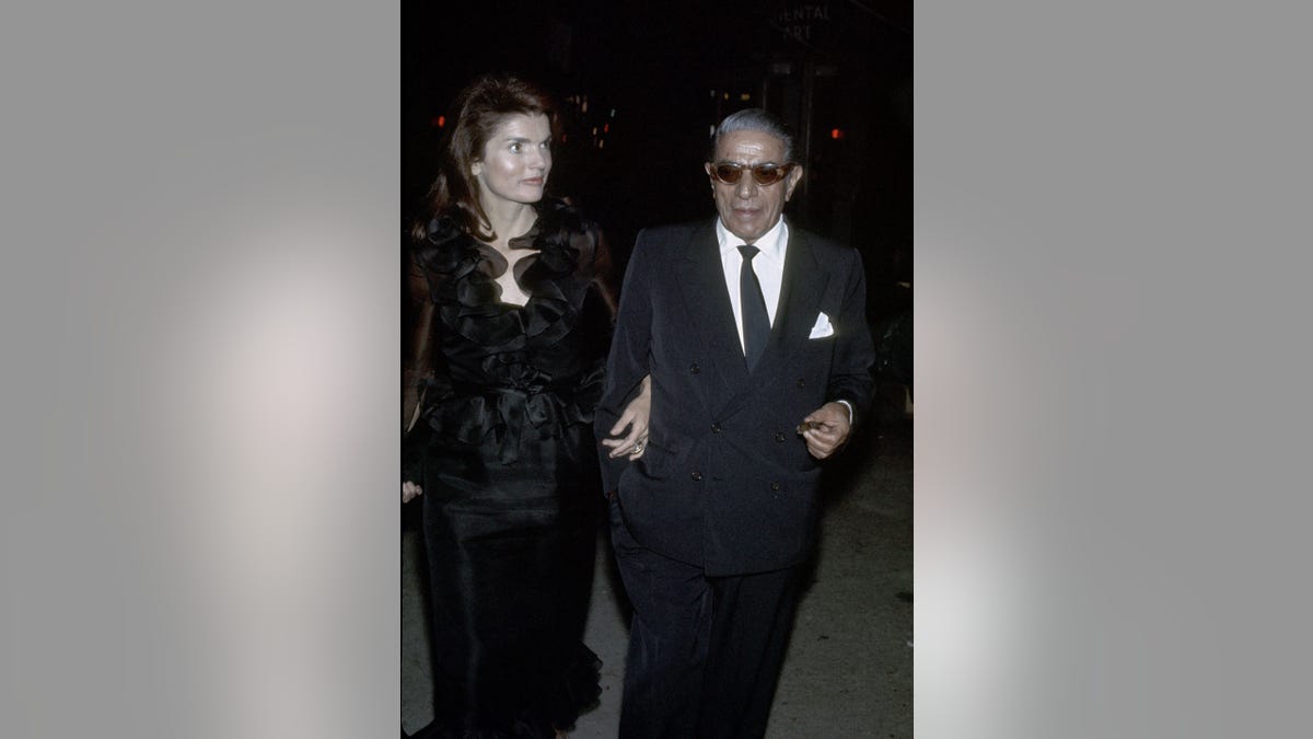 Jackie Kennedy wearing a coat in the arm of Onassis who was wearing a suit.