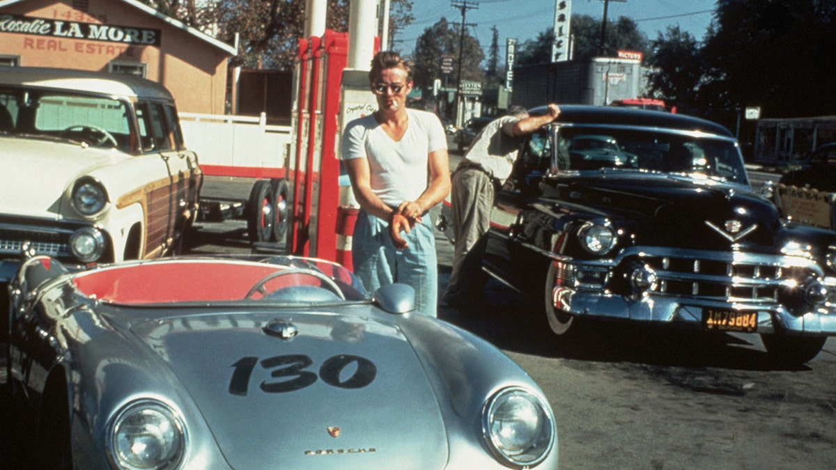 James Dean in a white shirt, blue jeans and sunglasses standing in front of his Porsche