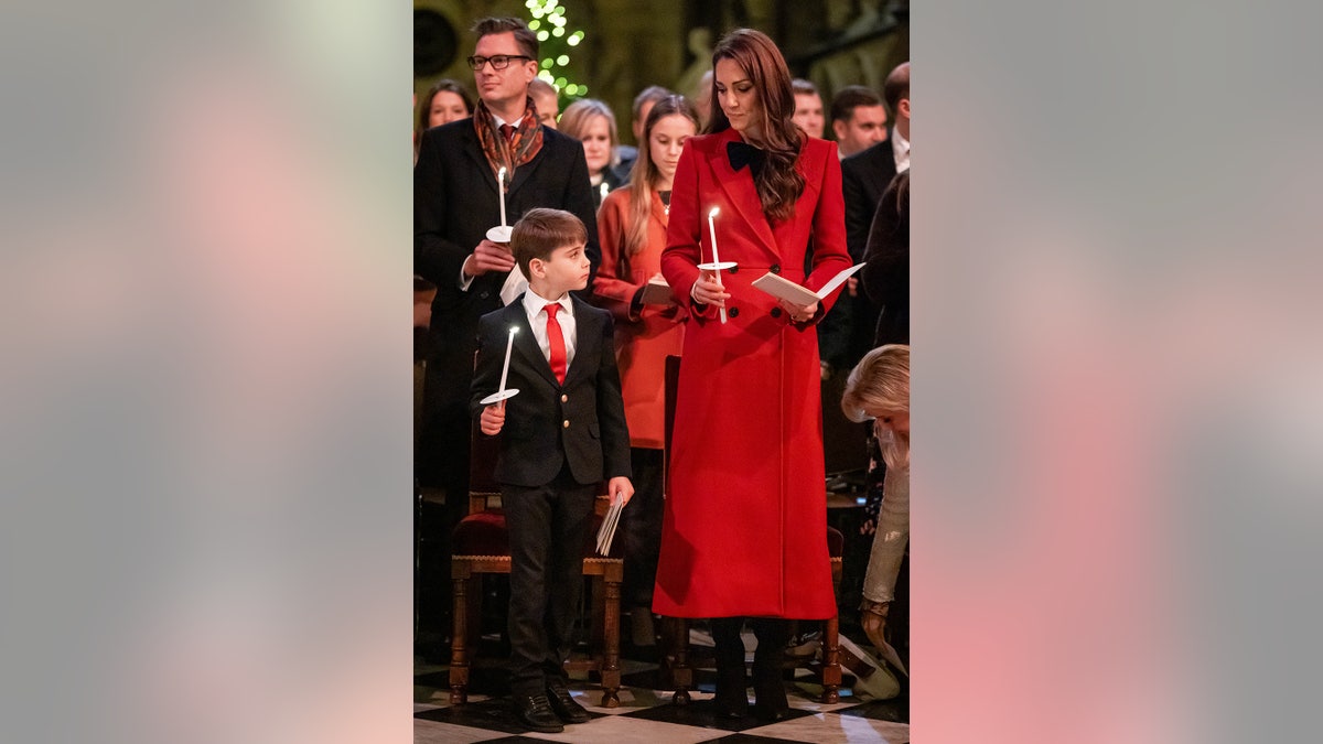 Kate Middleton in a red coat holding a candle and standing next to Prince Louis in a suit also holding a candle.