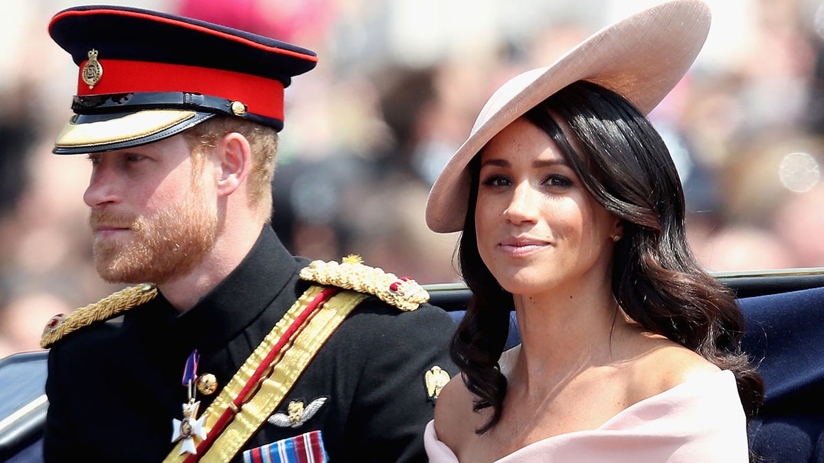 A close-up of Prince Harry in uniform and Meghan Markle in a pale pink dress with a matching hat sitting on a royal carriage.