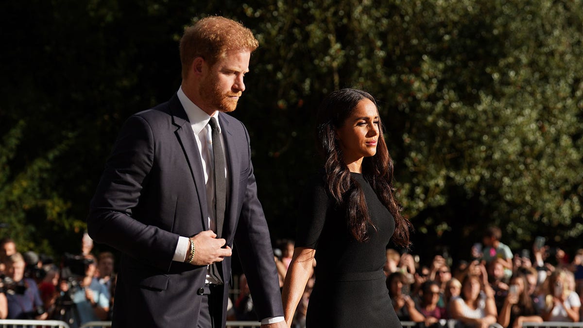 Prince Harry and Meghan Markle looking somber as they walk in public and hold hands wearing black.