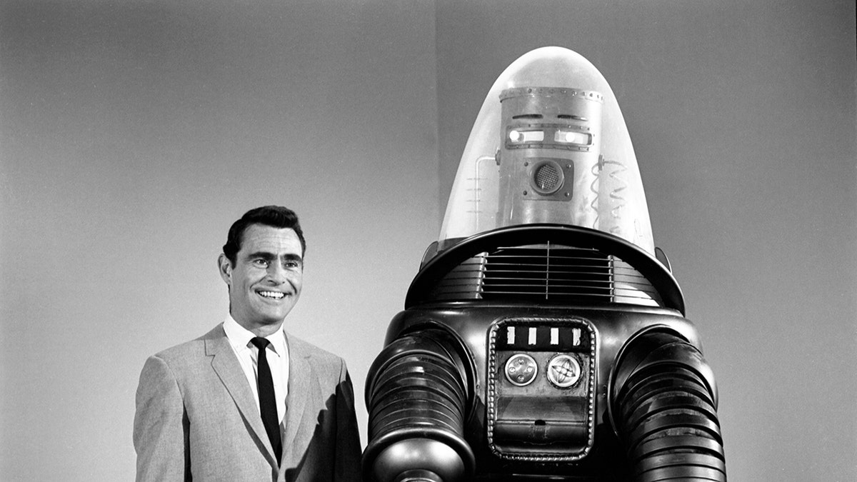 Rod Serling in a suit smiling alongside a robot.