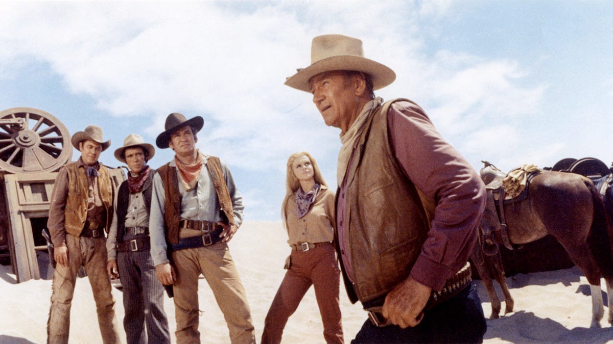 John Wayne looking to the side with several cowboys and a woman in Western gear.