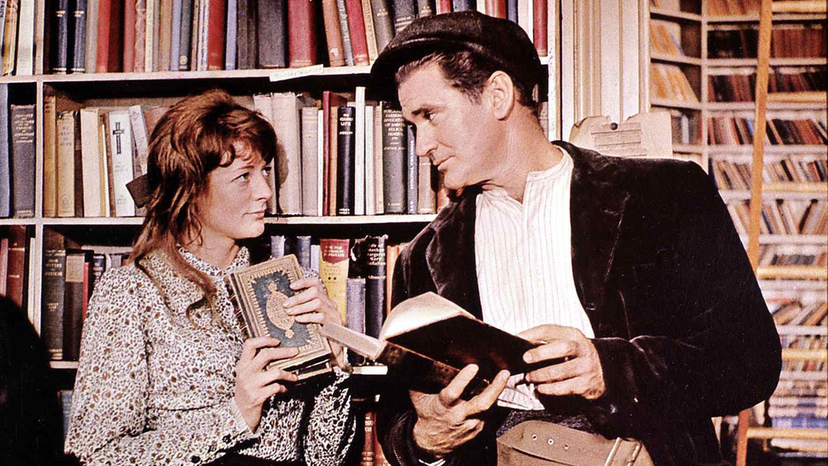 Maggie Smith and Rod Taylor holding books at a library as they smile at each other.