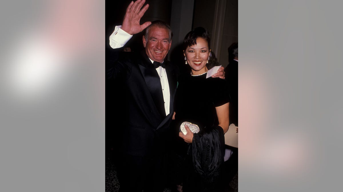 Rod Taylor in a tux smiling and waving with his arm around his wife.