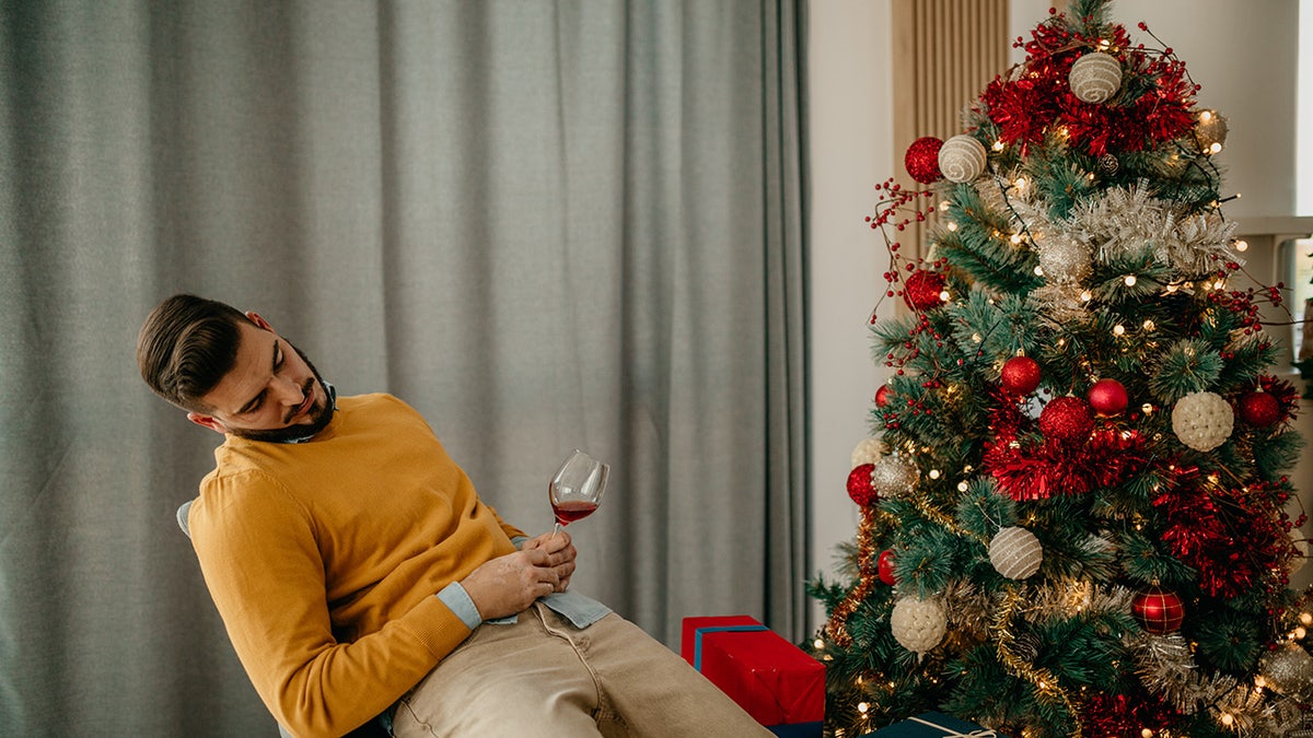 man sleeping on a chair next to the Christmas tree with Christmas gifts and a wine glass in his hands