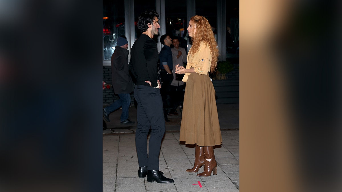 Justin Baldoni in a black shirt and navy pants talks with Blake Lively in an orange sweater and brown skirt on set of 'It Ends With Us'