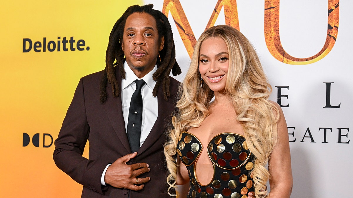 Jay-Z in a maroon jacket looks directly at the camera while wife Beyoncé in a gold circle dress smiles on the 'Mufasa: The Lion King' carpet