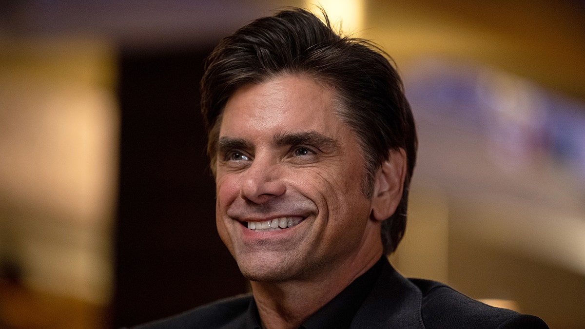 John Stamos grins and looks slightly up in a black jacket while being interviewed