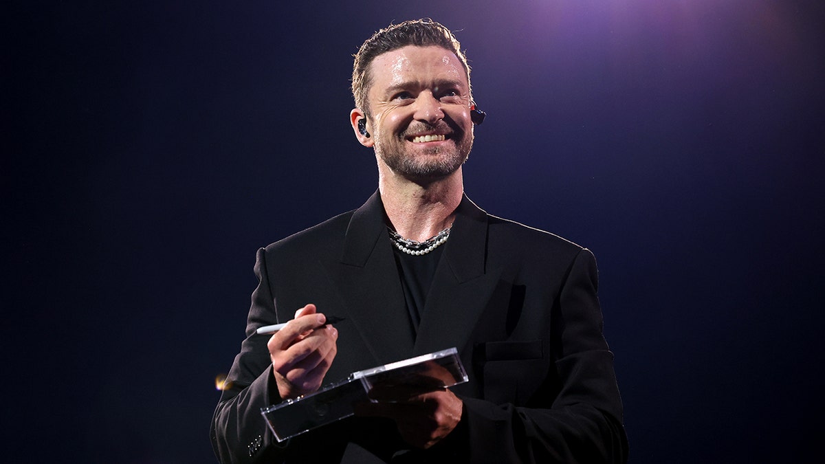 Justin Timberlake looks up gleefully on stage and smiles while signing a CD