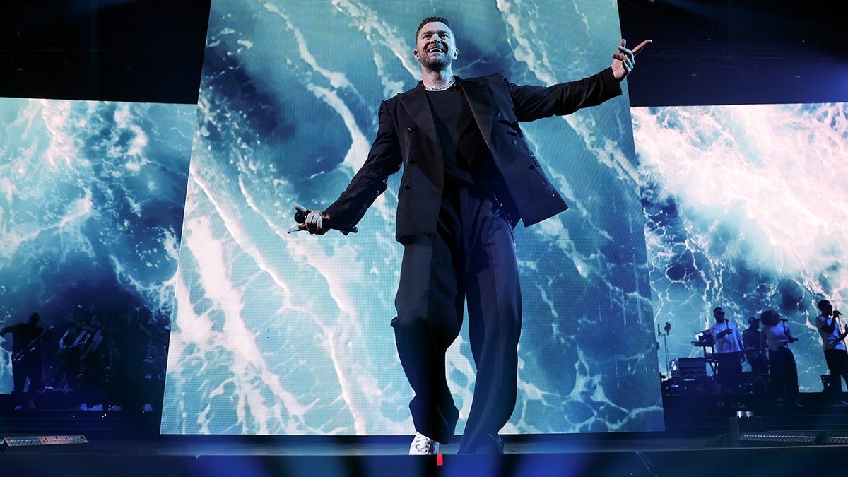 Justin Timberlake looks up as he stretches his arm out and performs on stage with a marble-like blue background