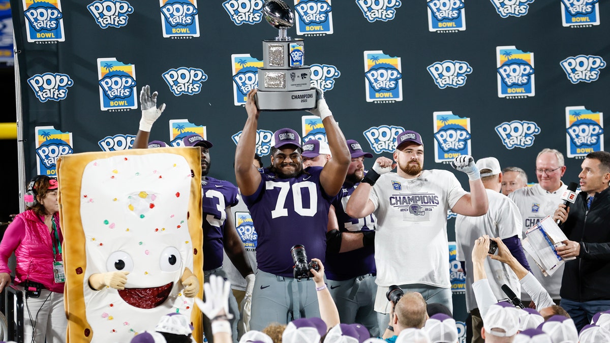 A Kansas State player hoists the Pop-Tarts Bowl trophy as his team celebrates with the game's mascot in 2023.