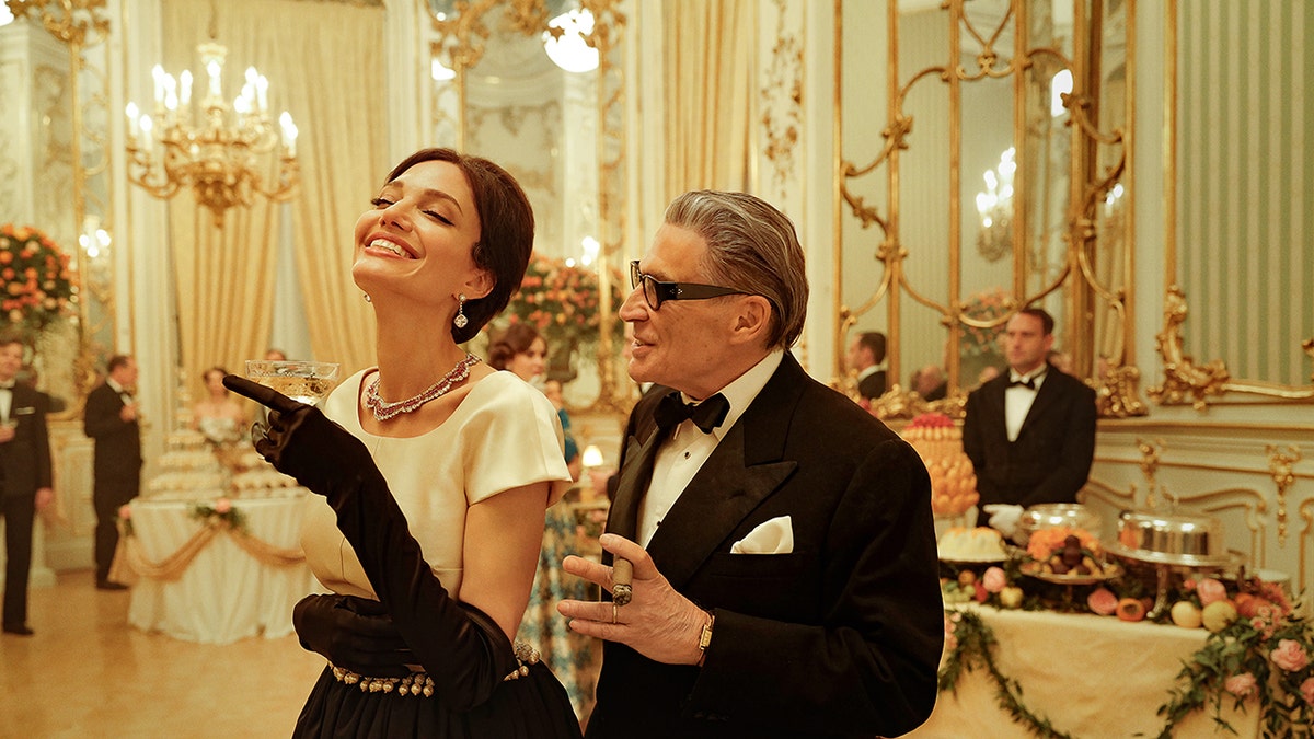 Angelina Jolie smiling as Maria Callas in a glamorous gown next to an actor in a suit playing Aristotle Onassis