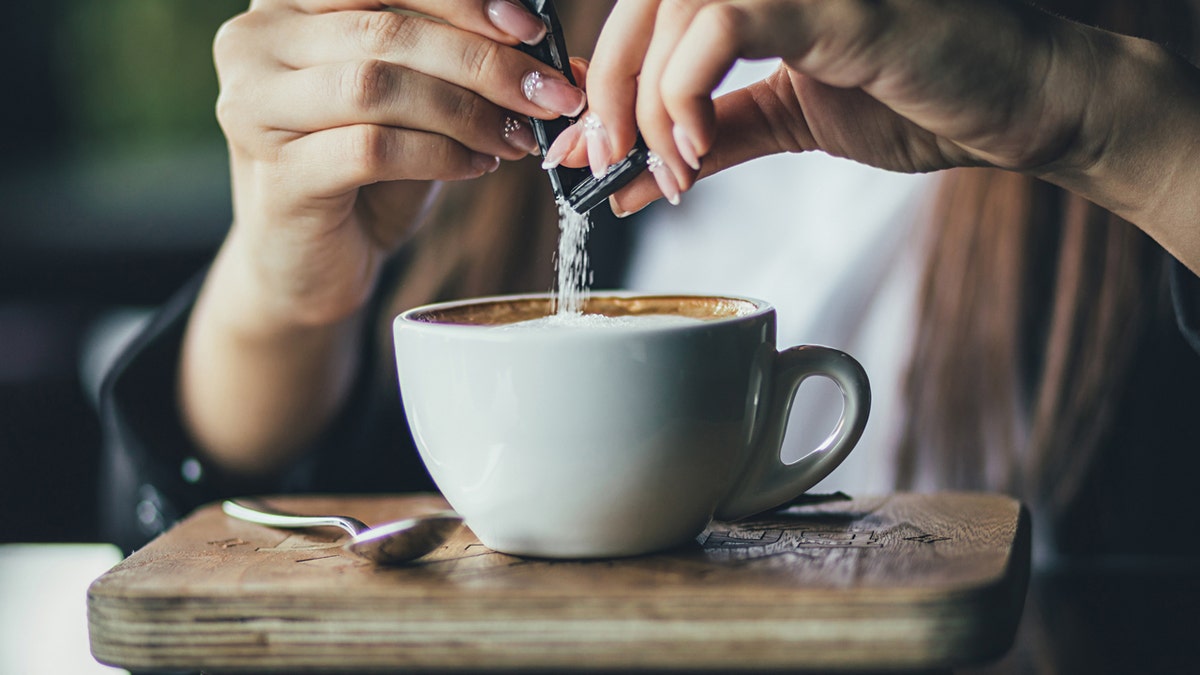 A woman's hands are pouring sugar from a packet into a cup of coffee.