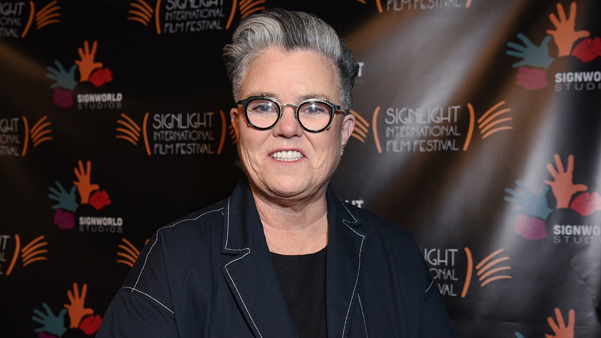 Rosie O'Donnell in a denim jacket smiles wearing circular black glasses on the carpet