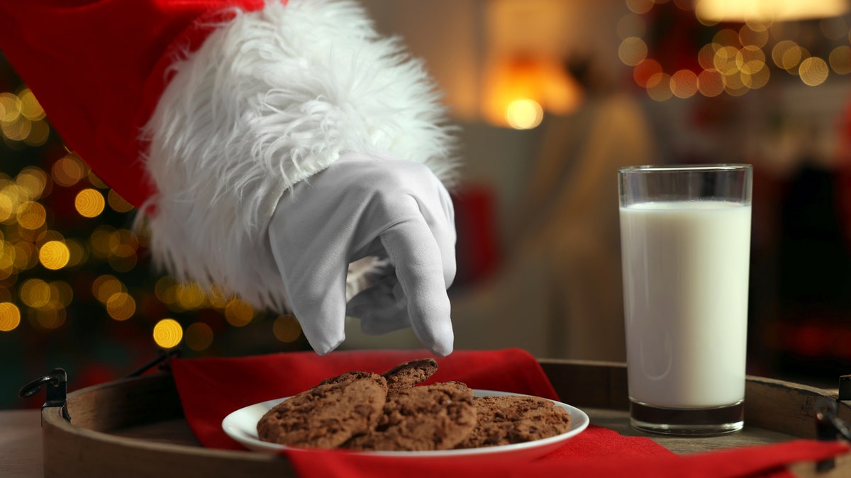 A hand belonging to Santa Claus reaches for some cookies on a plate next to a glass of milk.