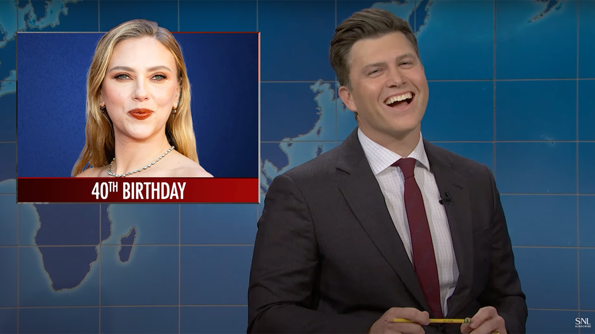 Colin Jost laughs in a grey suit and red tie behind the 