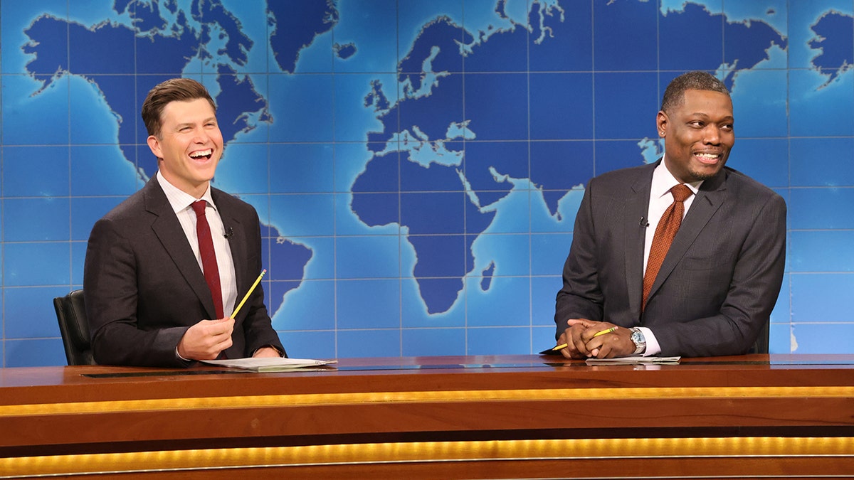 Colin Jost laughs out loud holding a pencil behind the 'Weekend Update' desk on 