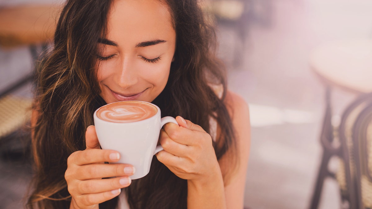 A woman smiles as she prepares to take a sip from a cup of cappuccino.