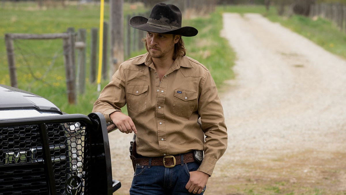 Luke Grimes in a black cowboy hat and tan shirt leans against a car in a scene from 