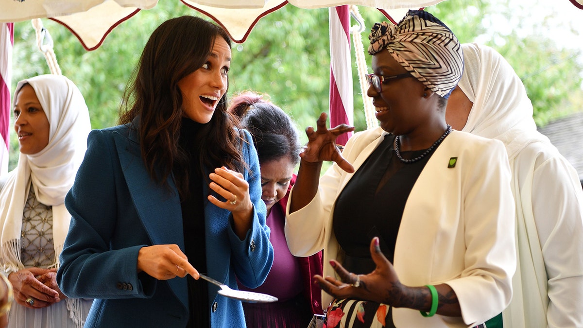 Meghan Markle speaking to a woman wearing a blue blazer and a black shirt.