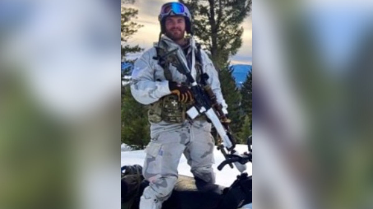 Matthew Livelsberger wearing cold weather gear and holding a rifle while standing over a snowmobile