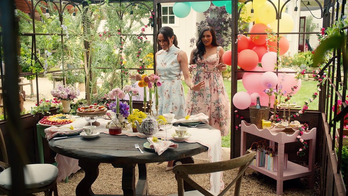 Mindy Kaling in a floral dress walking behind Meghan Markle heading toward a dining area.