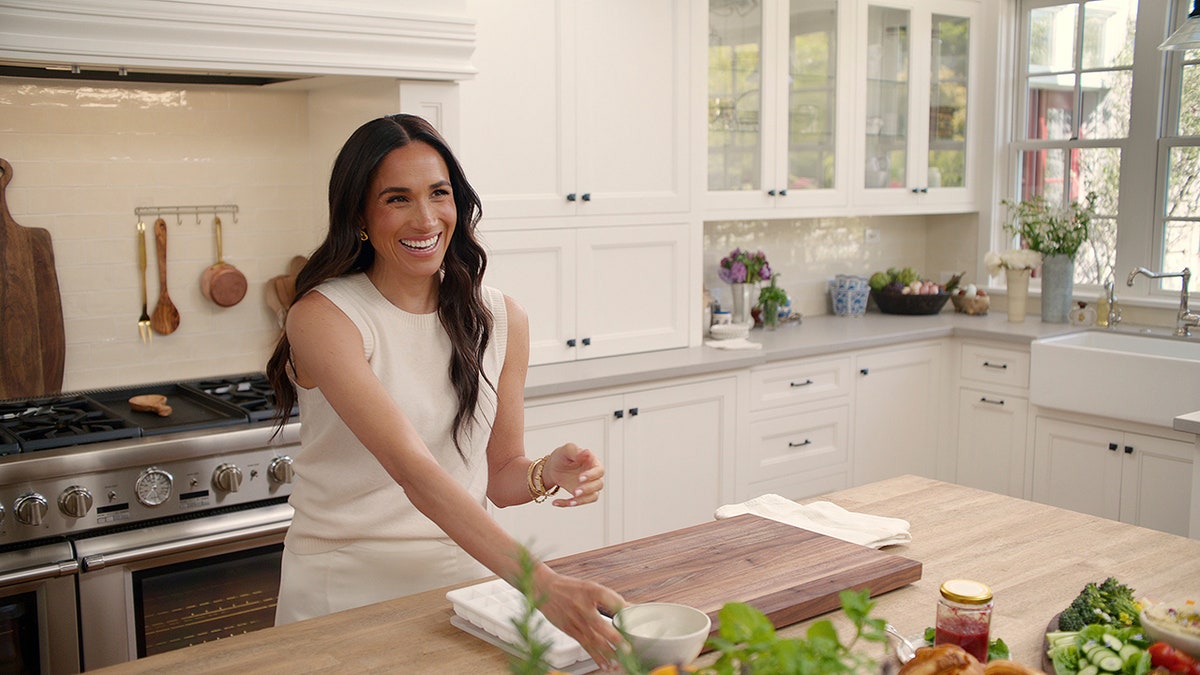 Meghan Markle wearing a creme sleeveless shirt inside a kitchen and smiling.