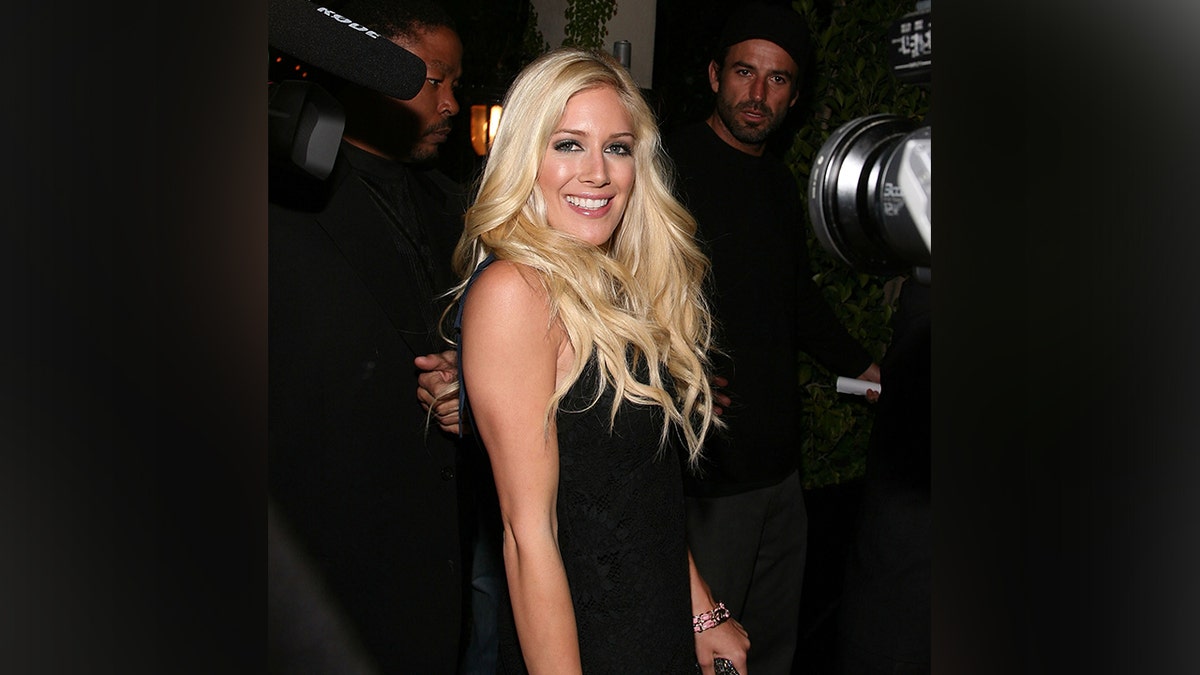 Heidi Montag in a black dress and platinum blonde hair smiles as she walks into her album release party